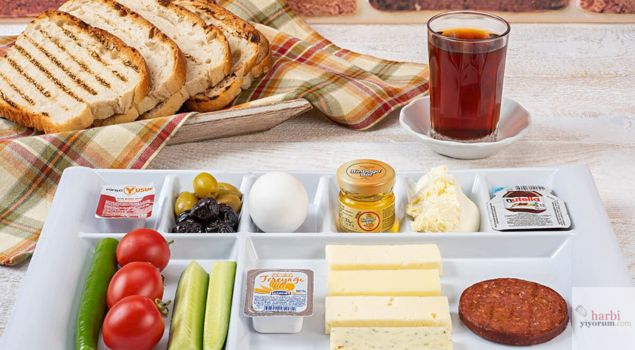 Bir kahvaltı tabağı görseli: Kızarmış ekmek dilimleri, bir bardak çay, zeytin çeşitleri, haşlanmış yumurta, bal, tereyağı, Nutella, taze domates, salatalık, yeşil biber, beyaz peynir, kaşar peyniri, çeşnili peynir ve sucuk dilimleri ile zengin bir kahvaltı sunumu.